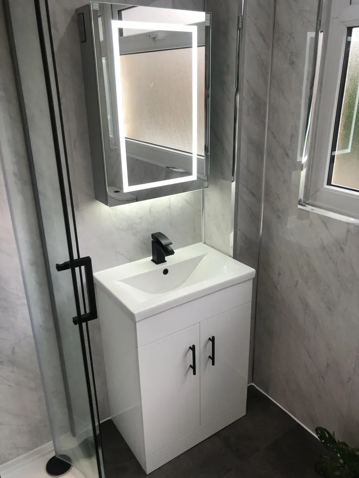 A white vanity unit under an illuminated mirror cabinet, with marble-effect panels